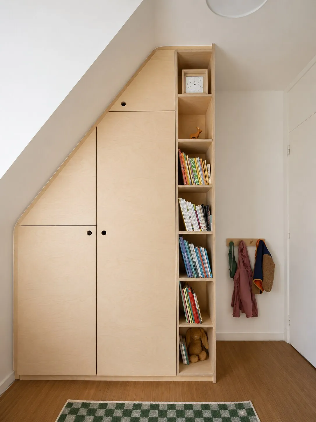 Hoge kast in berkenmultiplex met een schuin meegezaagde voorkant onder de dakhelling, met een smalle open boekenkolom aan de kamerzijde